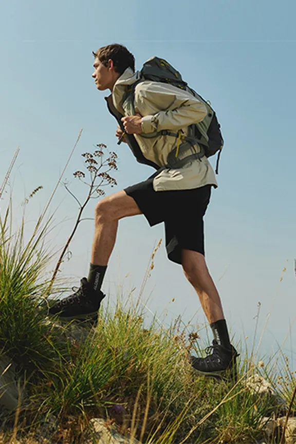 Ein Mann mit Rucksack wandert einen grasbewachsenen Hang unter blauem Himmel hinauf. 